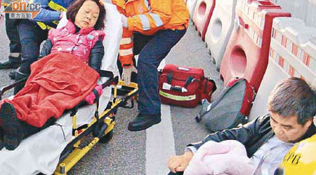 受傷婦由救護員送院，其丈夫傷勢較輕，坐在路邊。	（文健雄攝）