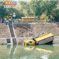 泥頭車由路面衝落河邊翻側，車頭半浸入水中。（陳錦昌攝）