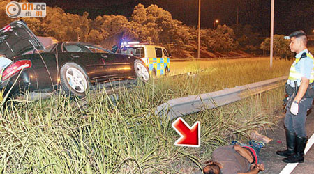 私家車剷上防撞欄，涉案南亞裔司機逃出車外昏倒路邊（箭嘴示）。（楊日權攝）