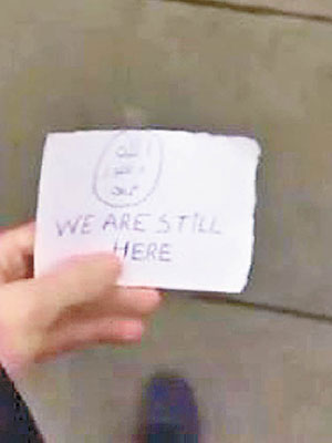一張寫有「我們還在這兒」（We are still here）的紙條及附近建築物，證明在安特衞普拍攝。（互聯網圖片）