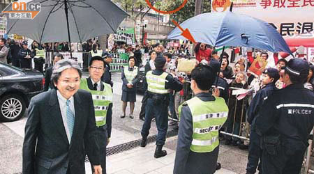 曾俊華入場時，有示威者（箭嘴示）向他投擲請願信（圓圈示），有保安為他打傘以防他被擲中。	（羅錦鴻攝）